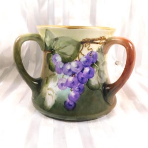 Puede incluir: Jarra de cerámica pintada a mano con dos asas. La jarra presenta un racimo de uvas moradas y hojas verdes. Las asas hacen la transición de verde a marrón. El borde está adornado con oro.