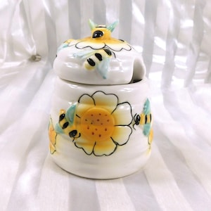 May include: A white ceramic honey pot with a lid, decorated with yellow sunflowers and black and yellow bees. The lid has a bee-shaped handle. The pot is designed for storing honey.