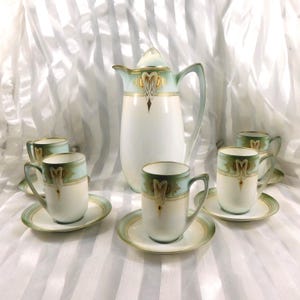 Op de afbeelding: Een vintage porseleinen theeservies met een hoge witte kan en bijpassende kopjes en schotels. De set heeft een lichtblauwe en gouden rand met decoratieve accenten. De kopjes hebben oren en staan op bijpassende schotels.
