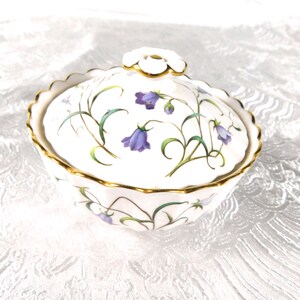 Könnte beinhalten: Eine weiße Zuckerdose aus Porzellan mit Deckel, verziert mit einem Blumenmuster aus violetten glockenförmigen Blüten und grünen Stielen. Die Dose und der Deckel haben einen goldfarbenen Wellenrand und einen blütenförmigen Griff am Deckel.