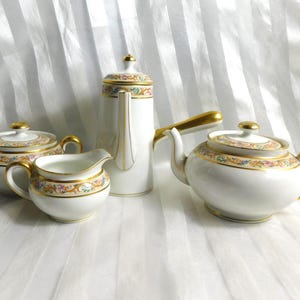 Puede incluir: Juego de té de porcelana blanca con ribetes dorados y detalles florales. El juego incluye una tetera, una lechera, una azucarera y una cafetera alta. Las asas y las tapas son doradas.