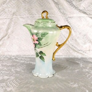 May include: A porcelain coffee pot featuring a soft green and white gradient finish with hand-painted pink flowers and green leaves. The vessel includes a gold-toned handle, a matching lid with a gold finial, and a scalloped base edge.