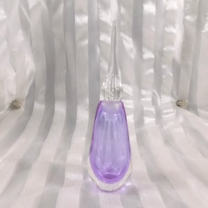 Può includere: Un flacone di profumo in vetro trasparente con interno lavanda e tappo allungato trasparente. Il flacone ha una forma a goccia con base arrotondata e lungo tappo a punta. Il vetro ha una finitura liscia e lucida.