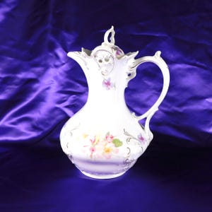 Può includere: Una brocca in porcellana bianca con coperchio, caratterizzata da delicati motivi floreali dipinti a mano in rosa e giallo, volute dorate e un manico curvo. Il recipiente presenta un bordo smerlato e un pomello decorativo sul coperchio.