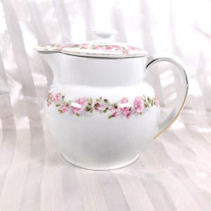 May include: White ceramic pitcher with a lid, adorned with a delicate pink floral garland around the body and lid. The handle and rim feature gold accents. The pitcher is designed for serving beverages.