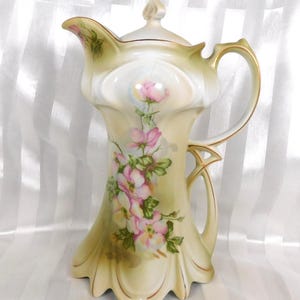 May include: A tall, antique-style ceramic pitcher with a lid. The pitcher is cream-colored with a floral design of pink roses and green leaves. Gold trim accents the edges and handle. The pitcher has a unique, flared shape.