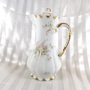 Pode incluir: Cafeteira de porcelana branca com tampa, com um design floral em rosa e verde. A cafeteira tem uma alça e acabamentos dourados. A cafeteira tem aproximadamente 25 cm de altura.