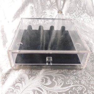 Może przedstawiać: Przezroczyste akrylowe pudełko na biżuterię z czarnym aksamitnym wnętrzem. Pudełko ma prostokątny kształt z otwieraną pokrywą i małą szufladą. Wnętrze posiada szczeliny na pierścionki i inne przedmioty biżuteryjne. Pudełko stoi na białej, wzorzystej powierzchni.