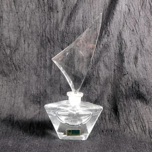 Peut inclure: Flacon de parfum en verre transparent avec un design géométrique. Le flacon a une base en forme de losange et un bouchon triangulaire. Une petite étiquette est apposée sur le devant. Le flacon est vide et posé sur une surface gris foncé.