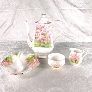 Op de afbeelding: Een wit porseleinen theeservies met een delicaat patroon van roze kersenbloesembomen. De set bevat een hoge theepot met deksel, een kleine melkkan, een suikerpot en een schaaltje met een geschulpte rand, allemaal afgewerkt met gouden accenten.