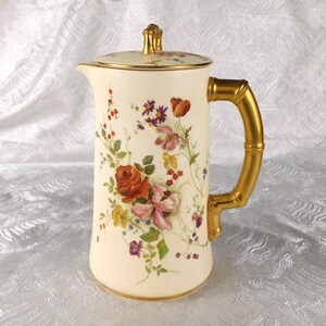 Puede incluir: Una cafetera de porcelana color crema con un arreglo floral pintado a mano con flores rojas, rosas y amarillas. El recipiente incluye un asa dorada con forma de bambú, un remate de tapa dorado a juego y un borde dorado.