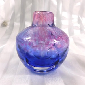 Puede incluir: Un jarrón de vidrio soplado a mano con un cuerpo redondeado y un cuello corto. El jarrón hace la transición de un azul profundo en la base a un patrón moteado rosa y púrpura en la parte superior. El vidrio tiene un acabado brillante.