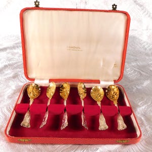 May include: A set of six gold-colored spoons in a red velvet-lined case. The spoons have ornate floral designs on the bowls and decorative handles. The case is red with a cream-colored interior and the text "I. MAGNIN" is visible.