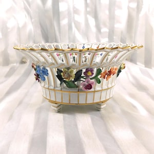 May include: A white porcelain bowl with a scalloped rim and floral detailing. The bowl has a lattice design, gold trim, and is supported by three small feet. It features colourful, three-dimensional flowers. The bowl is a decorative piece.