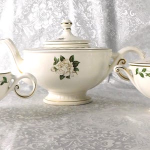 Könnte beinhalten: Cremefarbenes Teeservice mit einer Teekanne mit Rosenmotiv, einem Sahnekännchen und einer Zuckerdose mit Deckel. Das Set hat goldfarbene Verzierungen und grüne Blätter, die einen eleganten, klassischen Look erzeugen.