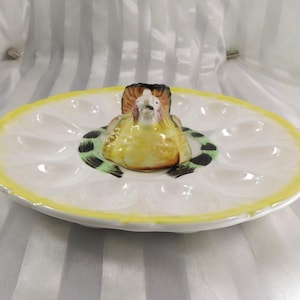 Op de afbeelding: Een vintage keramische eierserveerschaal met een gele rand en een centrale kippenfiguur. De witte schaal heeft eivormige inkepingen. De kip is geel, bruin en zwart en voegt een decoratief tintje toe aan de serveerschaal.