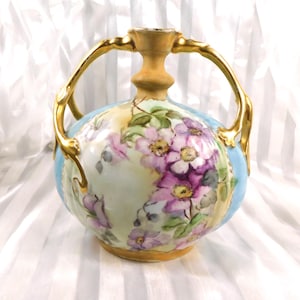 Op de afbeelding: Een decoratieve vaas met een bolvormig lichaam en twee goudkleurige handvatten. De vaas heeft een lichtblauwe achtergrond met handgeschilderde roze en paarse bloemmotieven. De vaas is ongeveer 25 cm hoog.