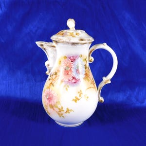 Może przedstawiać: Biały dzbanek do kawy z porcelany z ręcznie malowanym motywem kwiatowym przedstawiającym różowe, niebieskie i żółte kwiaty przypominające chryzantemy oraz zielone liście. Dzbanek posiada pasującą pokrywkę, wygięty uchwyt i złote wykończenia krawędzi.