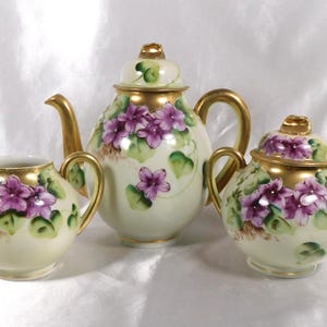 Puede incluir: Juego de té de porcelana de tres piezas, que incluye una tetera, una jarra para crema y un azucarero. Cada pieza está pintada con violetas moradas y hojas verdes, con detalles dorados. El juego se muestra sobre un fondo blanco.