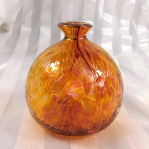May include: A hand-blown glass vase with a round body and a narrow neck. The vase is a blend of orange, yellow, and red hues, with a textured, swirling pattern throughout. The glass has a glossy finish, reflecting light.