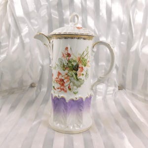 May include: A white porcelain coffee pot with a lid, featuring a floral design in shades of red, green, and peach. The pot has a purple ombre base and gold trim. The handle and spout are elegantly curved.