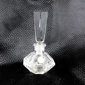 Peut inclure: Un flacon de parfum en verre clair et facetté, avec un bouchon rectangulaire et long. Le flacon a une base en forme de losange et un élément décoratif rond. Le fond est un tissu sombre avec un motif géométrique.