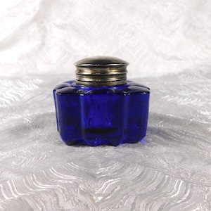 Pode incluir: Um tinteiro de vidro azul cobalto com tampa de metal prateado. O tinteiro tem uma forma canelada, semelhante a uma flor. A tampa é redonda e tem um design canelado. O tinteiro está sobre uma superfície branca texturizada.