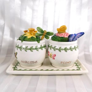 May include: A white ceramic set featuring two lidded jars and a rectangular tray. The jars are decorated with floral and insect motifs, including a dragonfly and butterfly. The jars are labelled "NARCISSUS" and "PEONY".