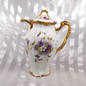 May include: A white porcelain coffee pot with a lid, adorned with purple and white floral designs and gold trim. The handle and spout are also gold. The pot has a fluted body and a decorative handle on the lid.