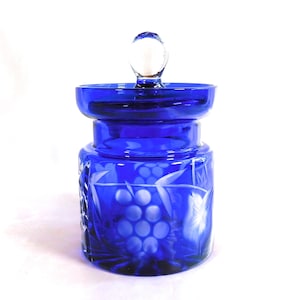 Puede incluir: Tarro de cristal azul cobalto con un pomo de cristal transparente en la tapa. El tarro presenta un diseño de cristal tallado de uvas y hojas. El tarro es una pieza decorativa, probablemente para guardar o exhibir.