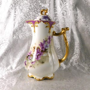 May include: White porcelain pitcher with a lid, adorned with purple floral designs and gold accents. The handle and trim are gilded. The pitcher has a bulbous body and a decorative lid with a small handle. A classic piece for serving beverages.
