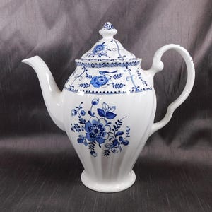 Pode incluir: Bule de porcelana branco com um design floral azul. O bule apresenta um bico curvo, uma alça grande e uma tampa com um botão decorativo. O padrão floral azul é repetido na tampa e no corpo do bule.