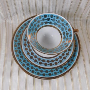 May include: A vintage teacup, saucer, and dessert plate set. The set features a light blue background with a repeating gold floral pattern. The rims are trimmed with gold. The teacup is white inside.