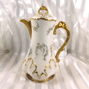Może przedstawiać: Biały porcelanowy dzbanek do kawy ze złotymi akcentami. Dzbanek ma zakrzywioną wylewkę, ozdobny uchwyt i pokrywkę ze złotym guzikiem. Delikatne kwiatowe wzory w pastelowych kolorach zdobią korpus, a złote obramowania podkreślają krawędzie.