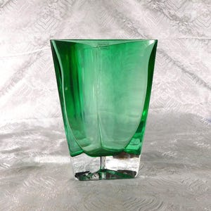 May include: A vibrant green glass vase with a unique, angular design. The vase features a clear, rectangular base that supports the green, curved upper portion. The glass has a glossy finish, reflecting light. The vase is suitable for floral arrangements.