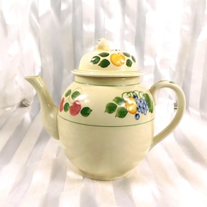 Pode incluir: Bule de cerâmica creme com tampa, decorado com ilustrações coloridas de frutas. O bule apresenta um bico e uma alça curvos, e uma linha verde circunda o corpo. A tampa é encimada por um pequeno botão decorativo.