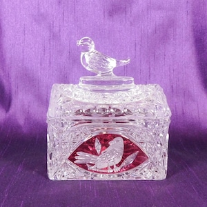 Puede incluir: Una caja de baratijas de vidrio transparente, de forma cuadrada, con una tapa en forma de pájaro. La caja presenta un diseño ovalado rojo con un pájaro grabado en su interior. El vidrio tiene un patrón tallado en diamante.