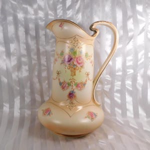 May include: A vintage cream-coloured pitcher with a curved handle and flared spout. It features a floral design with pink, purple, and blue roses. Gold trim adorns the edges and handle, providing an elegant touch to the piece.