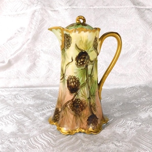 May include: A porcelain jug featuring a hand-painted design of brown pinecones and green pine needles against a soft cream and peach background. The vessel includes a gold-trimmed handle, a scalloped base, and a matching lid with a gold finial.