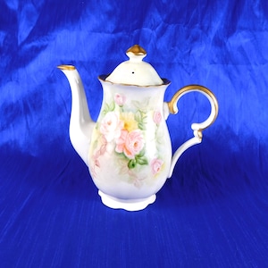 Puede incluir: Una tetera de porcelana blanca con un diseño floral pintado a mano de rosas rosas y amarillas, hojas verdes y bordes dorados en el asa, el pico y el remate de la tapa. La tetera tiene una base festoneada y una tapa extraíble a juego.