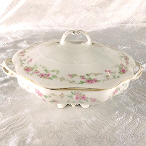 May include: A white porcelain serving dish with a lid, adorned with pink floral patterns and gold trim. The dish has two handles and sits on small feet. The lid features a decorative handle.