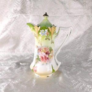 Può includere: Brocca vintage in porcellana con coperchio, con un design floreale di rose rosa e altri fiori. La brocca ha un manico bianco e finiture verdi e dorate. Il coperchio è verde con un pomello nero. Oggetto decorativo.
