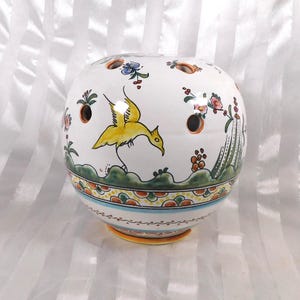 May include: A white ceramic flower frog with a round shape and multiple holes. The frog is decorated with a yellow bird, floral designs, and a green, orange, and blue border. The base is orange.