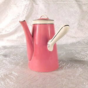 May include: A pink ceramic teapot with a white lid and a white handle. The teapot features a tall, slender spout and a smooth, glossy finish. A thin black line accents the rim of the lid and the edge of the handle, providing a clean contrast.
