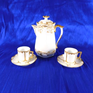 Puede incluir: Una cafetera de porcelana blanca con tapa y dos tazas a juego con sus platos. Cada pieza presenta detalles dorados ornamentados en los bordes, las asas y la base. La cafetera tiene un asa curva y un pico vertedor para servir el café.