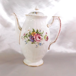 Puede incluir: Cafetera de porcelana blanca con diseño floral. La cafetera tiene tapa, boquilla y asa con detalles rojos. El diseño floral incluye flores rosas, moradas, amarillas y azules. Los bordes están adornados con ribetes dorados.