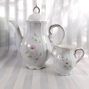 May include: A white porcelain tea set featuring a teapot with a lid and a creamer. The set is decorated with pink floral designs and gold trim. The teapot has a curved spout and handle. The creamer has a small handle and spout.