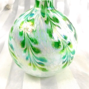 May include: A round, clear glass vase with a wide base and narrow neck. The vase is decorated with a pattern of green and white swirls, creating a leaf-like design. The glass has an iridescent quality, reflecting light.