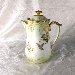 May include: An antique porcelain coffee pot with a lid, featuring a pale green body and gold trim. The pot is adorned with hand-painted floral and butterfly motifs. The handle and lid finial are also accented with gold.