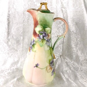 Puede incluir: Una jarra de porcelana pintada a mano con tapa. La jarra presenta un diseño floral de violetas moradas y hojas verdes sobre un fondo color crema. El asa y la tapa están adornadas con ribetes dorados. La jarra mide aproximadamente 30 cm de altura.